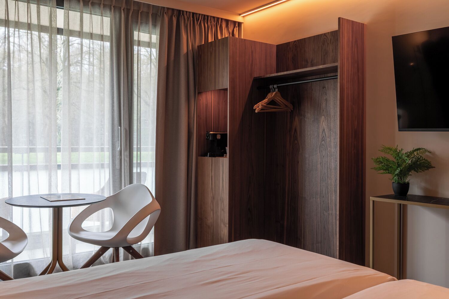 A hotel room with a bed, a small table with chairs, a coat rack, a potted plant, and a TV on the wall.