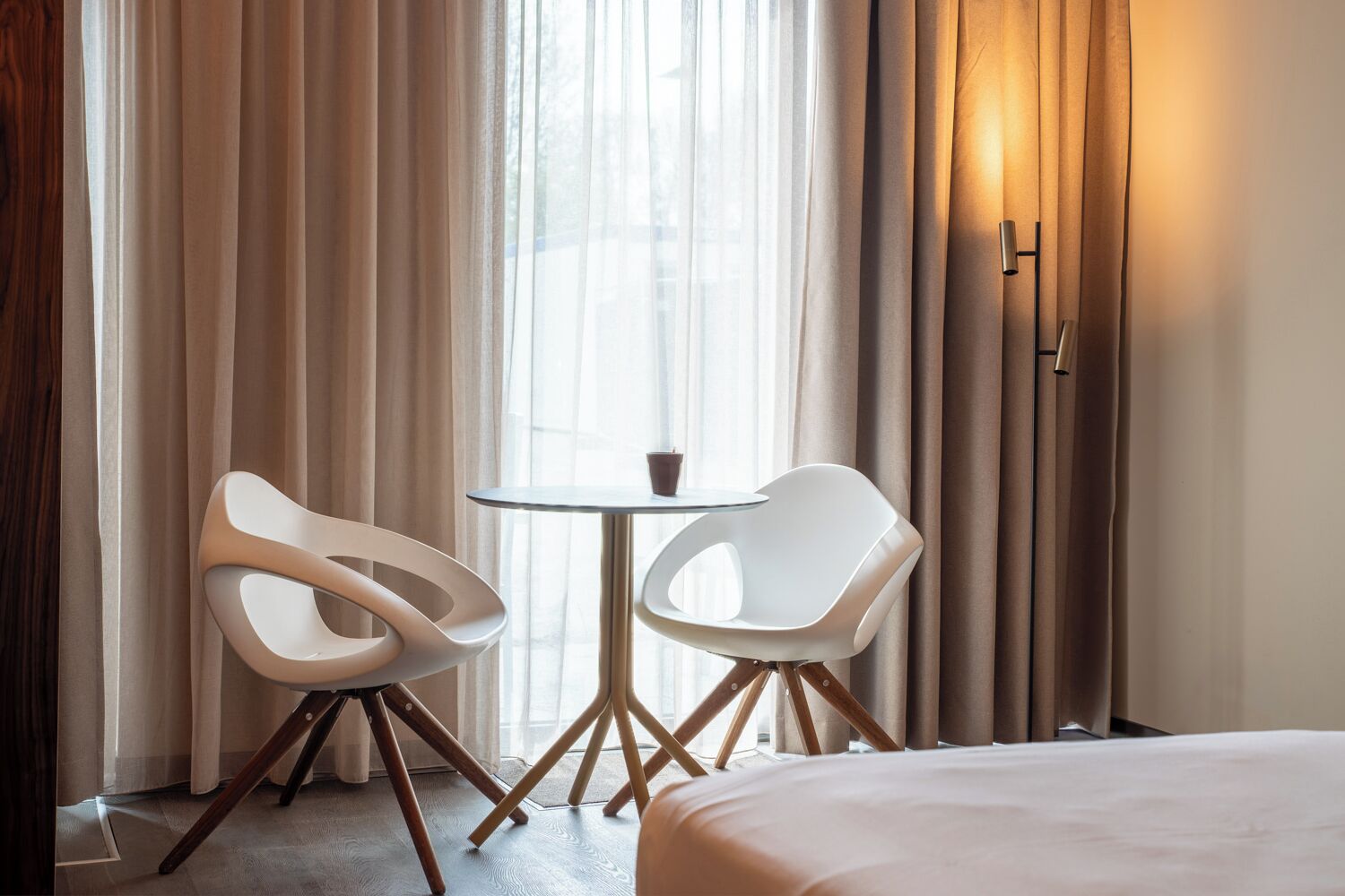 Een hotelkamer met een bed, twee witte stoelen, een klein rond tafeltje en een raam met gordijnen.