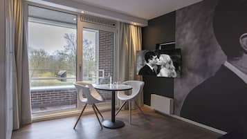 Une chambre moderne dotée d’une grande fenêtre, d’une table ronde avec deux chaises et d’un téléviseur fixé au mur, sur lequel on peut voir un couple.