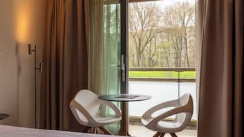 A hotel room with a bed, two white chairs, a small table, and a view of trees and a body of water through a glass door.