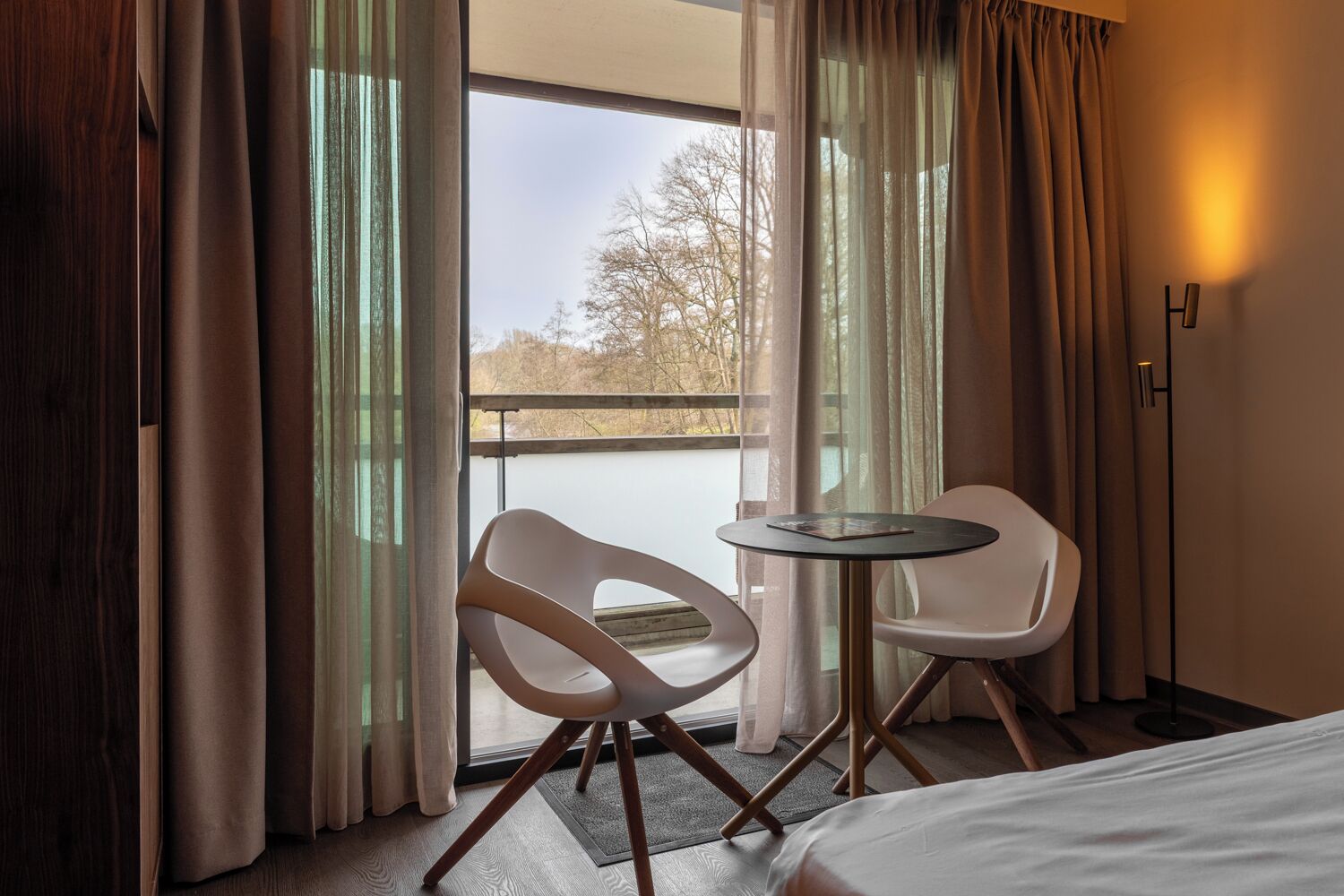 Habitación de hotel con cama, dos sillas blancas, una mesita y vistas a un lago a través de amplias ventanas.