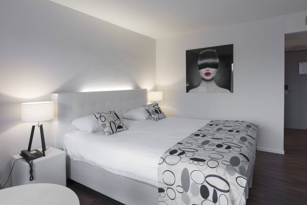A modern hotel room with a neatly made bed, a bedside lamp, a phone on the nightstand, and a large abstract painting on the wall.