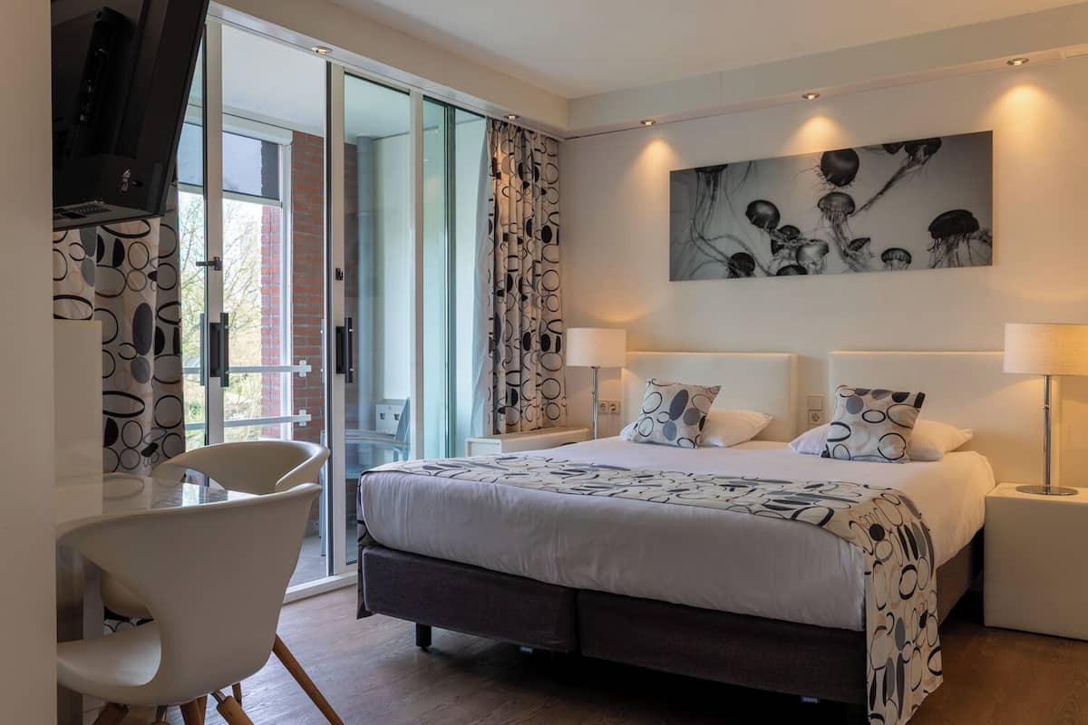 A modern hotel room with a large bed, a desk, and a glass door leading to a balcony.