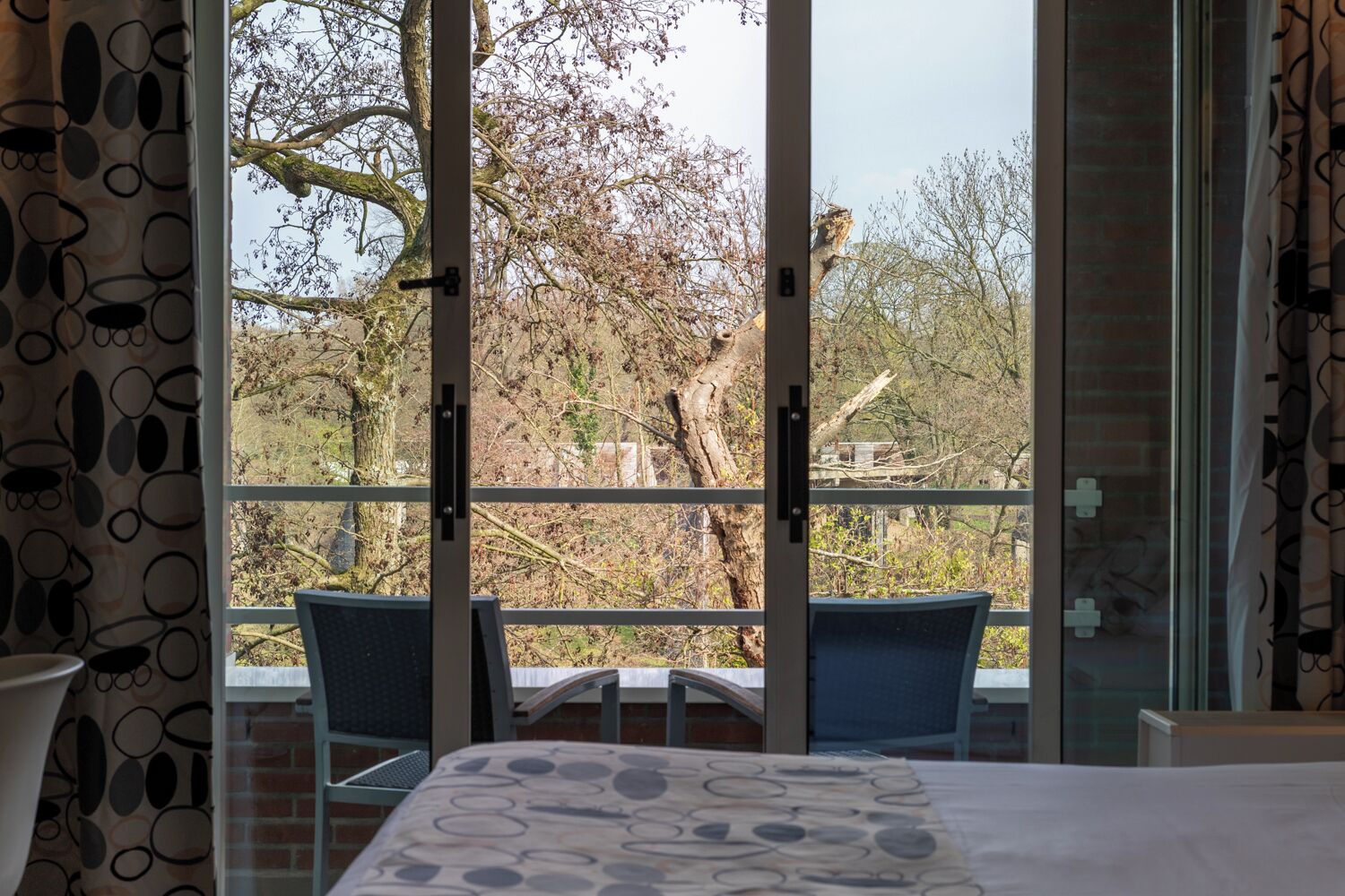 Een slaapkamer met een bed, een raam met uitzicht op bomen en een balkon met stoelen.