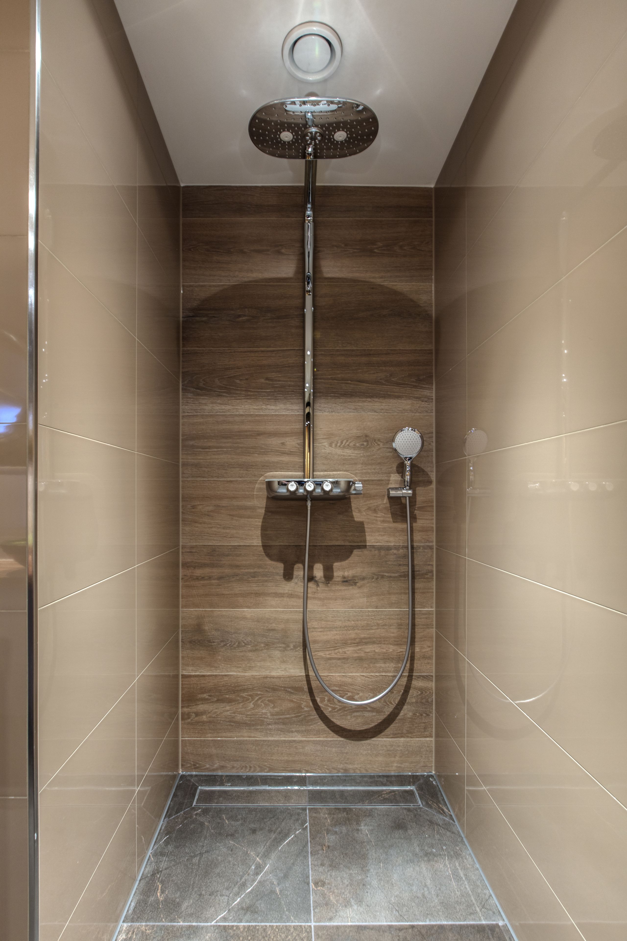 A modern shower with a handheld showerhead and a fixed overhead showerhead, set against a wooden wall with a tiled floor.