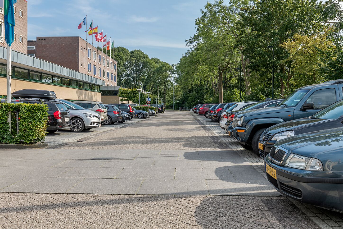 A parking lot with cars parked in rows, a building with multiple flags, and trees in the background.