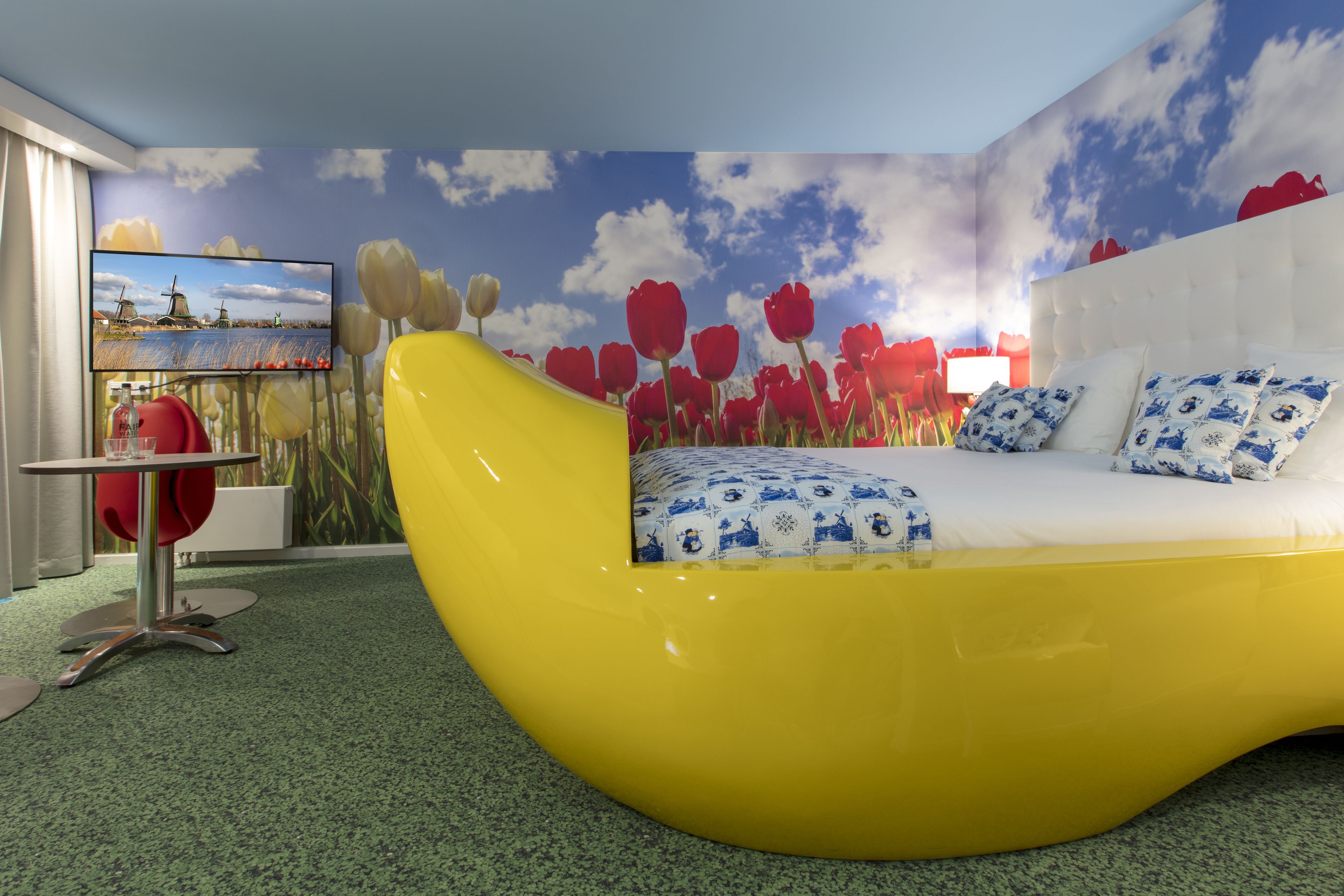 A modern bedroom with a curved yellow bed, a large TV on the wall, and a mural of tulips and a landscape.