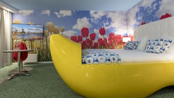 A modern bedroom with a curved yellow bed, a large TV on the wall, and a mural of tulips and a landscape.