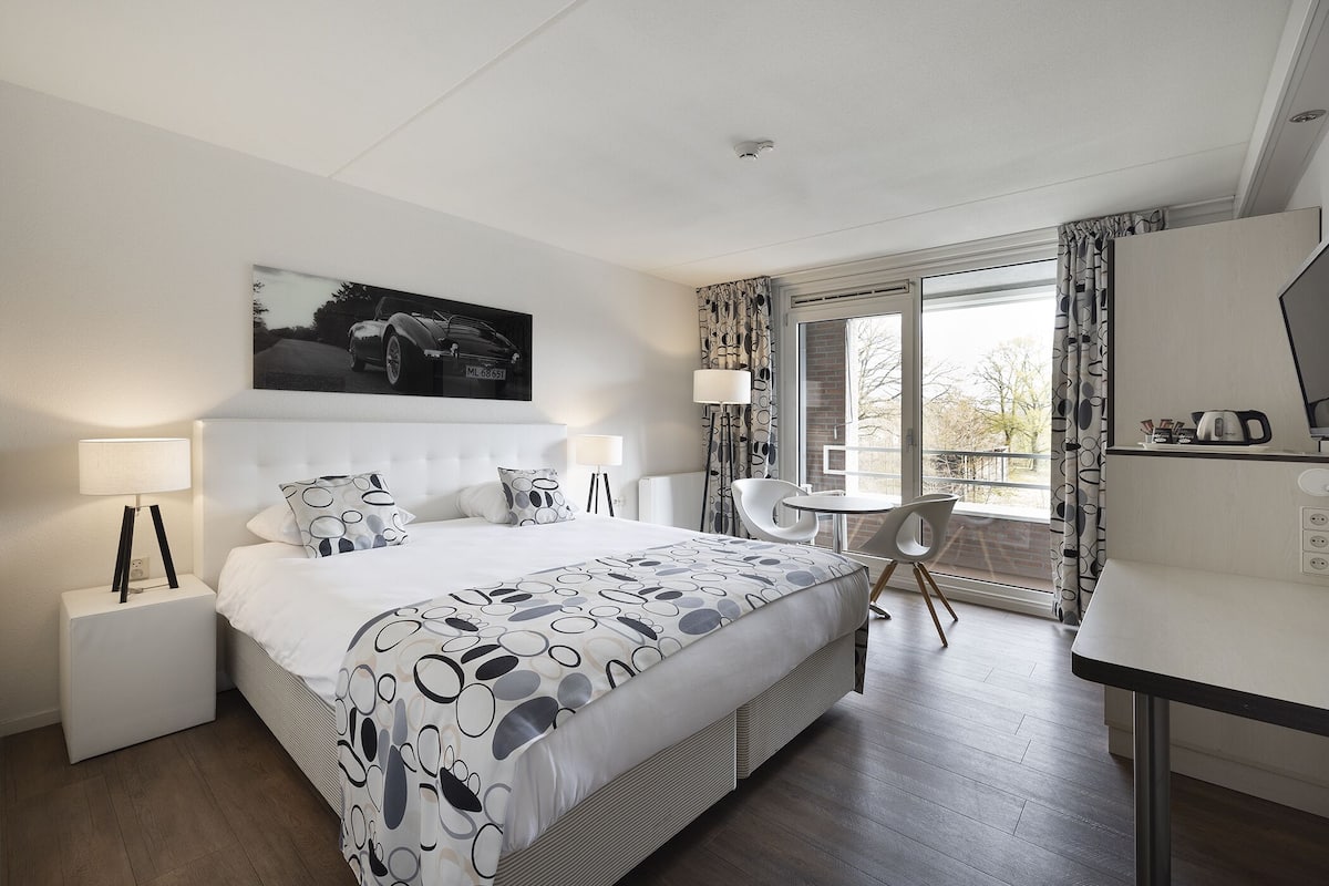 A modern hotel room with a large bed, a desk, and a balcony with curtains.