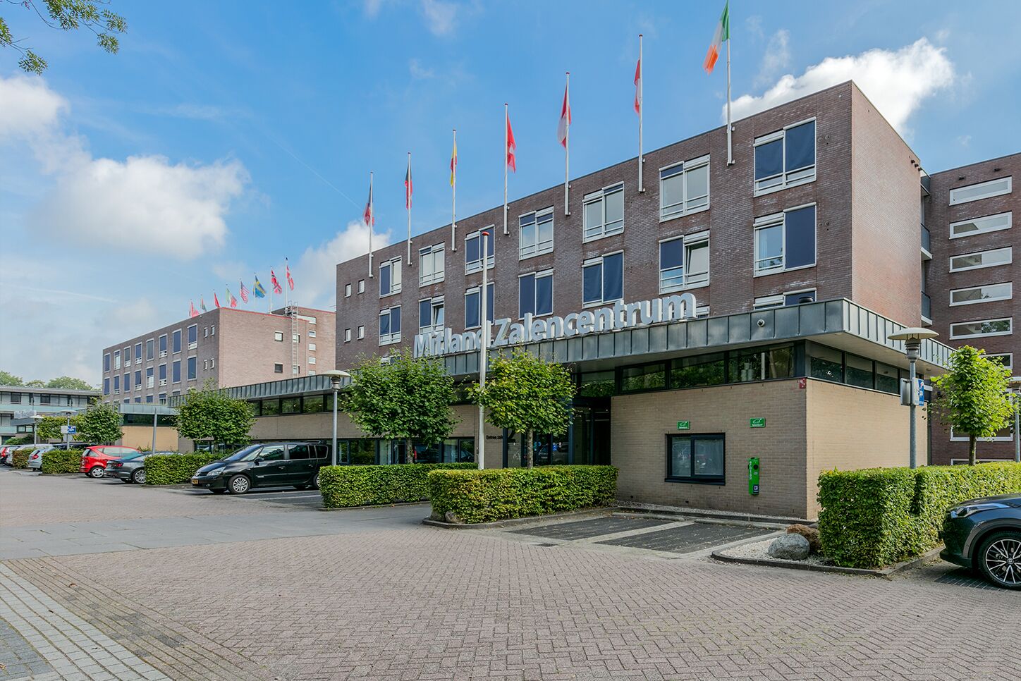 Un edificio moderno con un letrero que dice 'Milland Zaalcentrum'. Hay varias banderas en el techo, un estacionamiento con coches y un jardín bien cuidado.