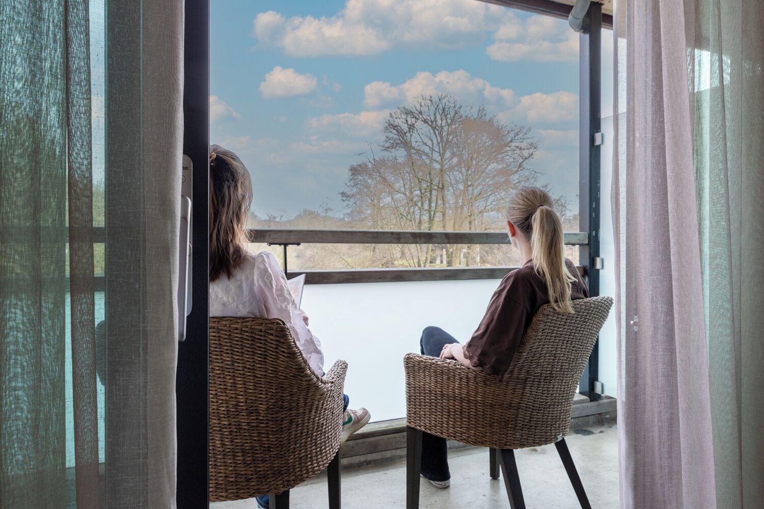 Två flätade stolar står placerade framför en balkong med utsikt över träd och en klar himmel.