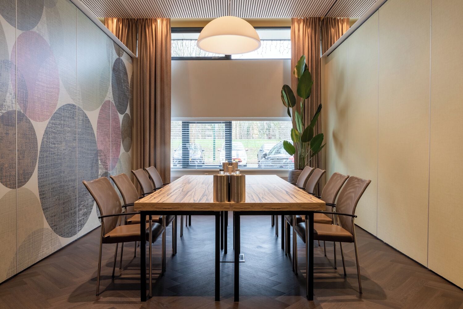 Un salón de conferencias con una mesa larga, sillas, una planta grande y una ventana con persianas.