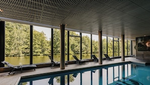 Una piscina interior con vistas despejadas a un lago y árboles, rodeada de sillones de descanso y amplias ventanas.