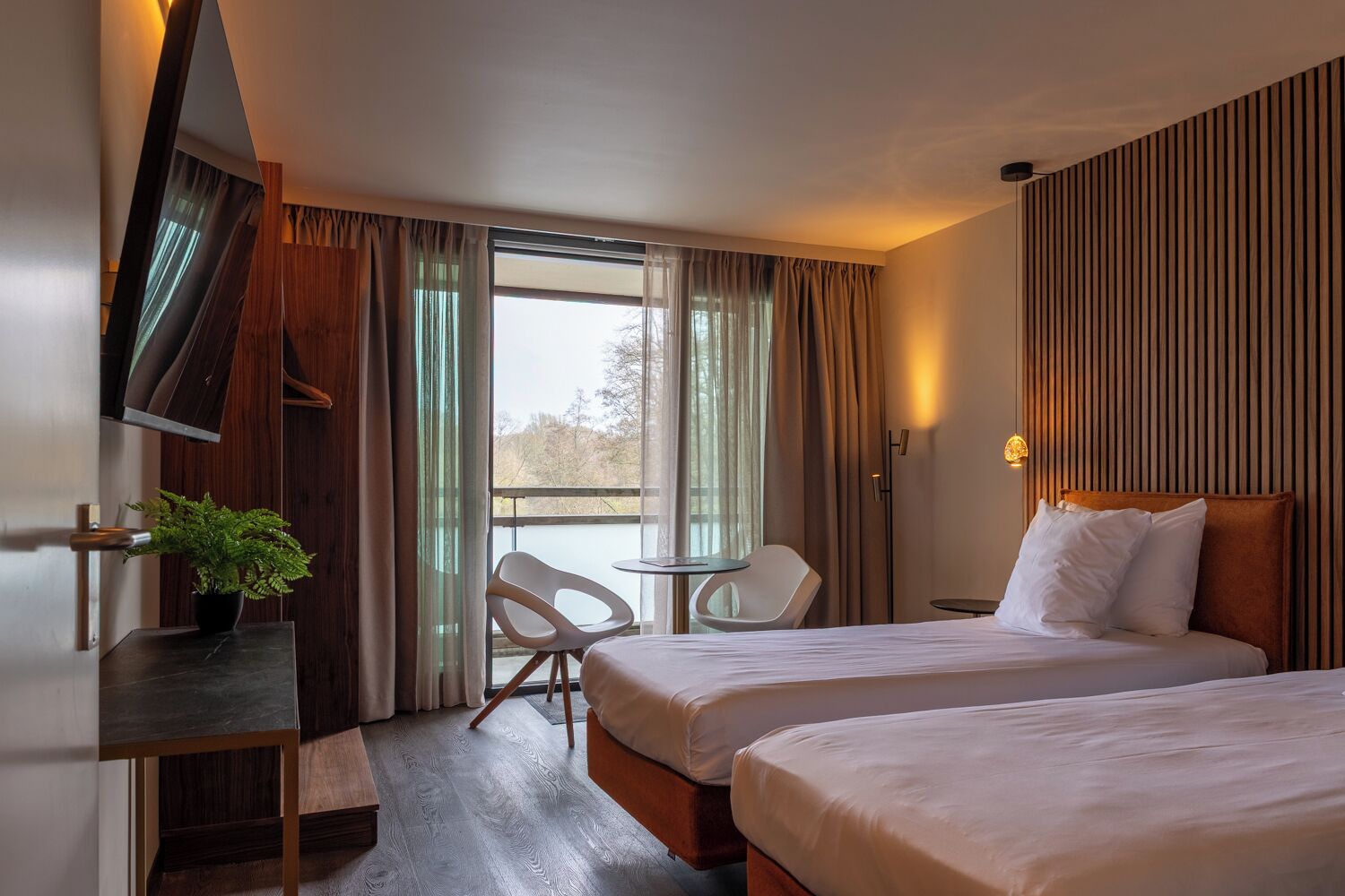 A hotel room with two beds, a desk, a chair, a potted plant, and a view of the outdoors.