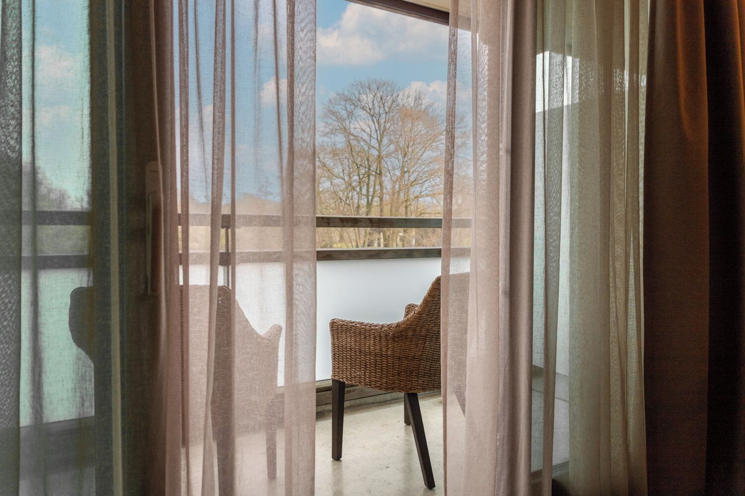 Una habitación con vistas, cortinas translúcidas, una silla de mimbre y un balcón con barandilla.