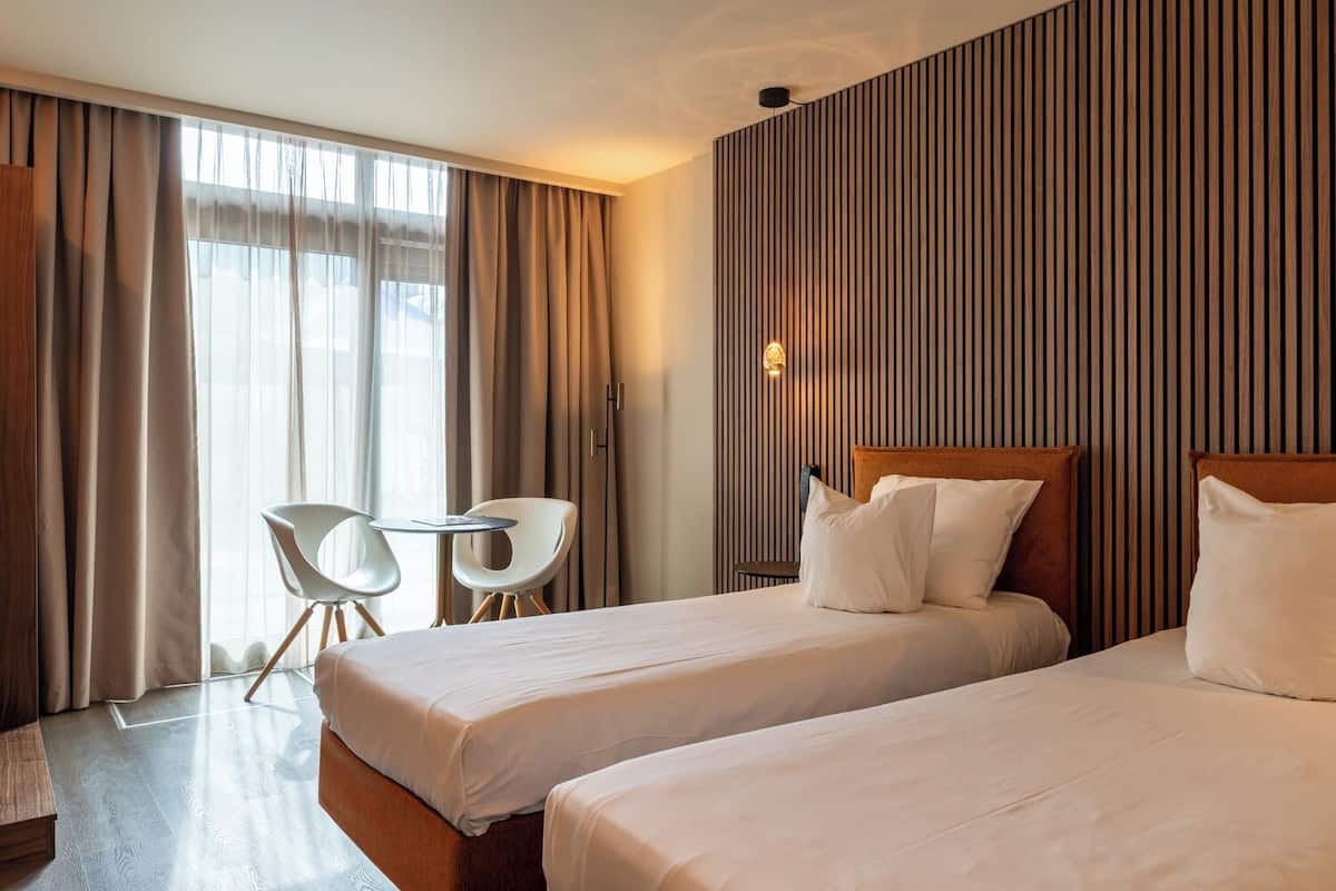 A hotel room with two beds, a small table, two chairs, and a large window with curtains.
