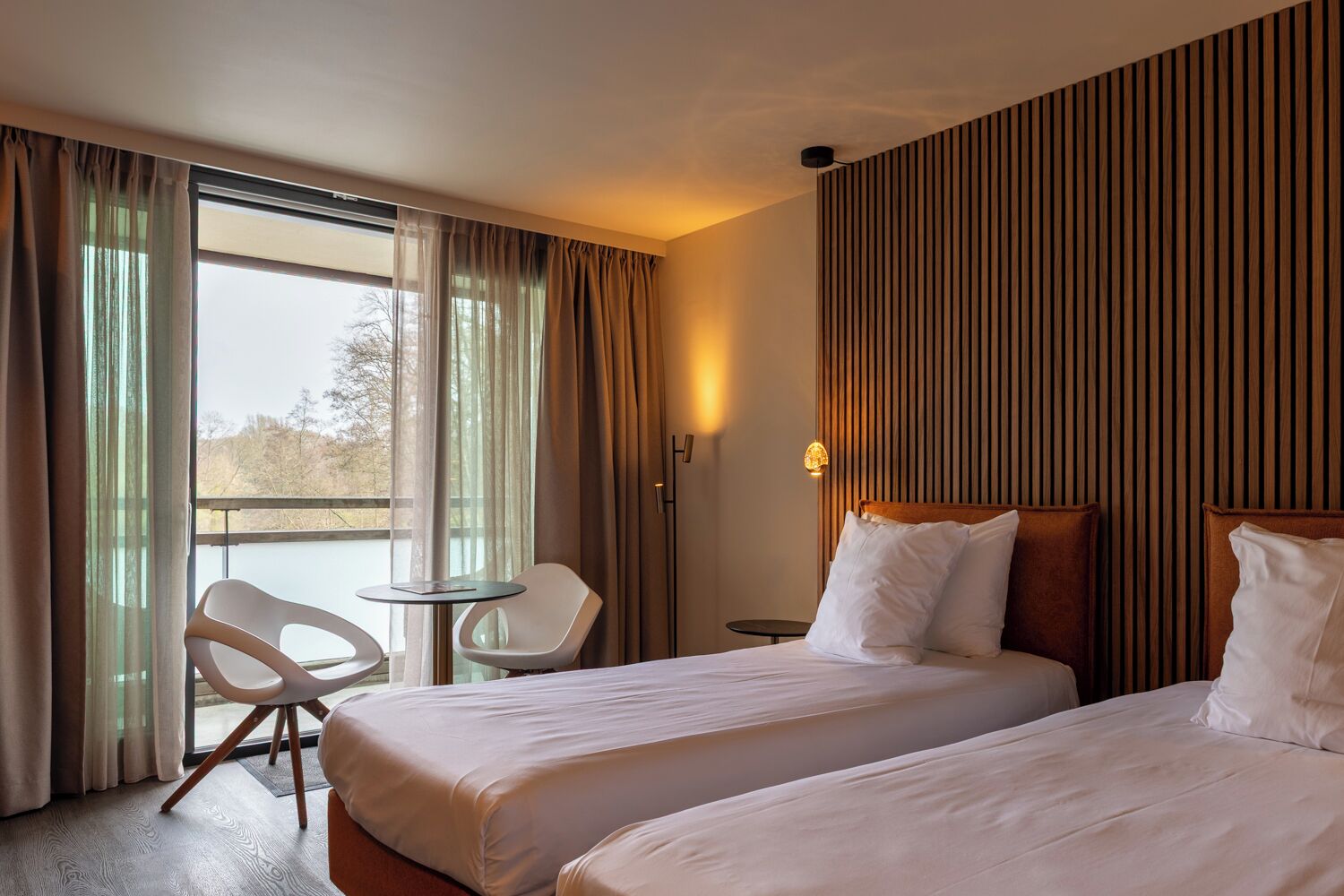 A hotel room with two beds, a chair, a small table, and a view of a body of water through large windows.