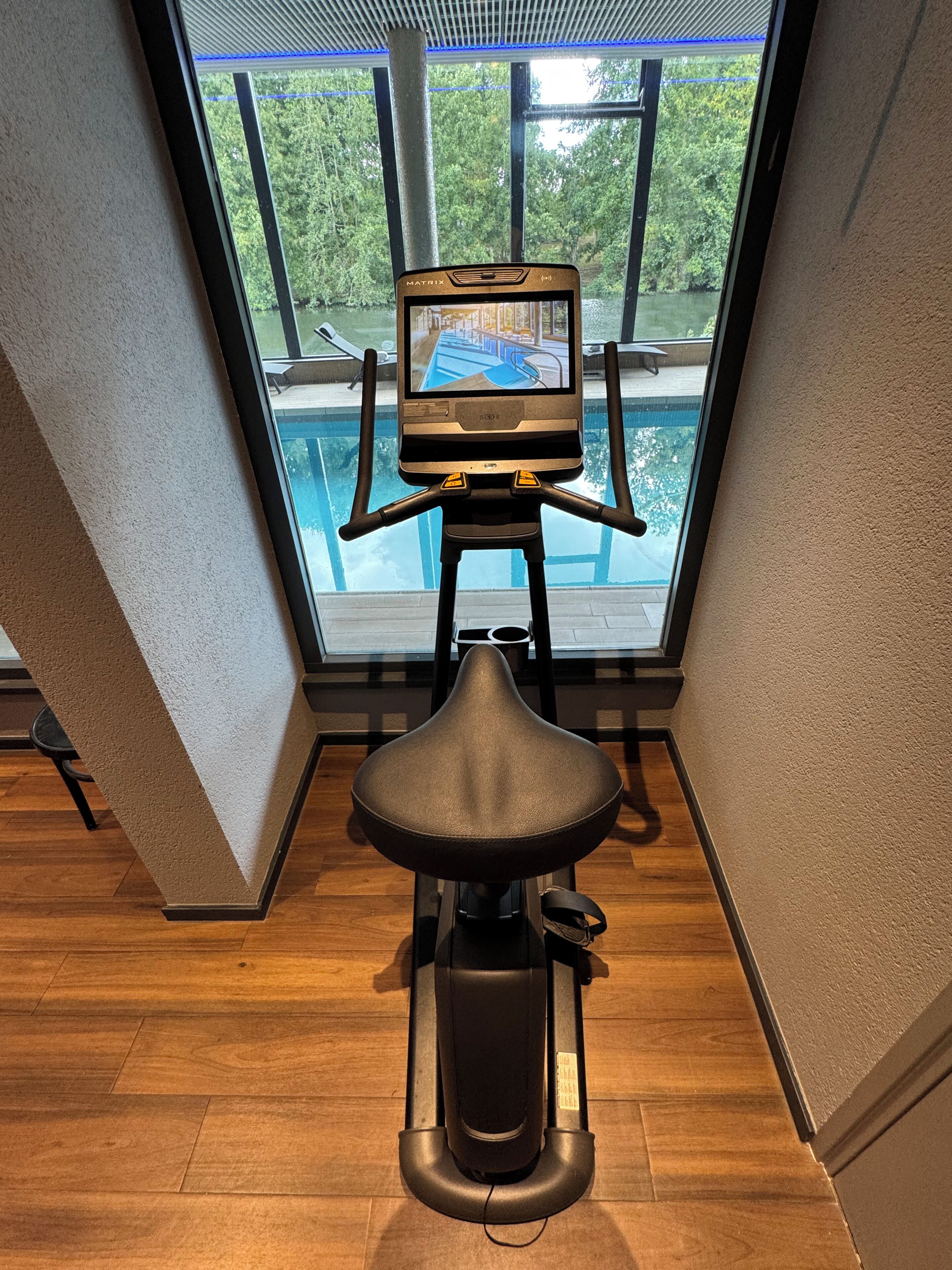 A stationary exercise bike with a screen, positioned in a room with a view of a swimming pool and greenery outside.
