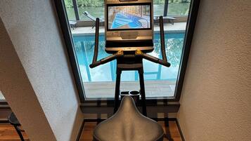 A stationary exercise bike with a screen, positioned in a room with a view of a swimming pool and greenery outside.