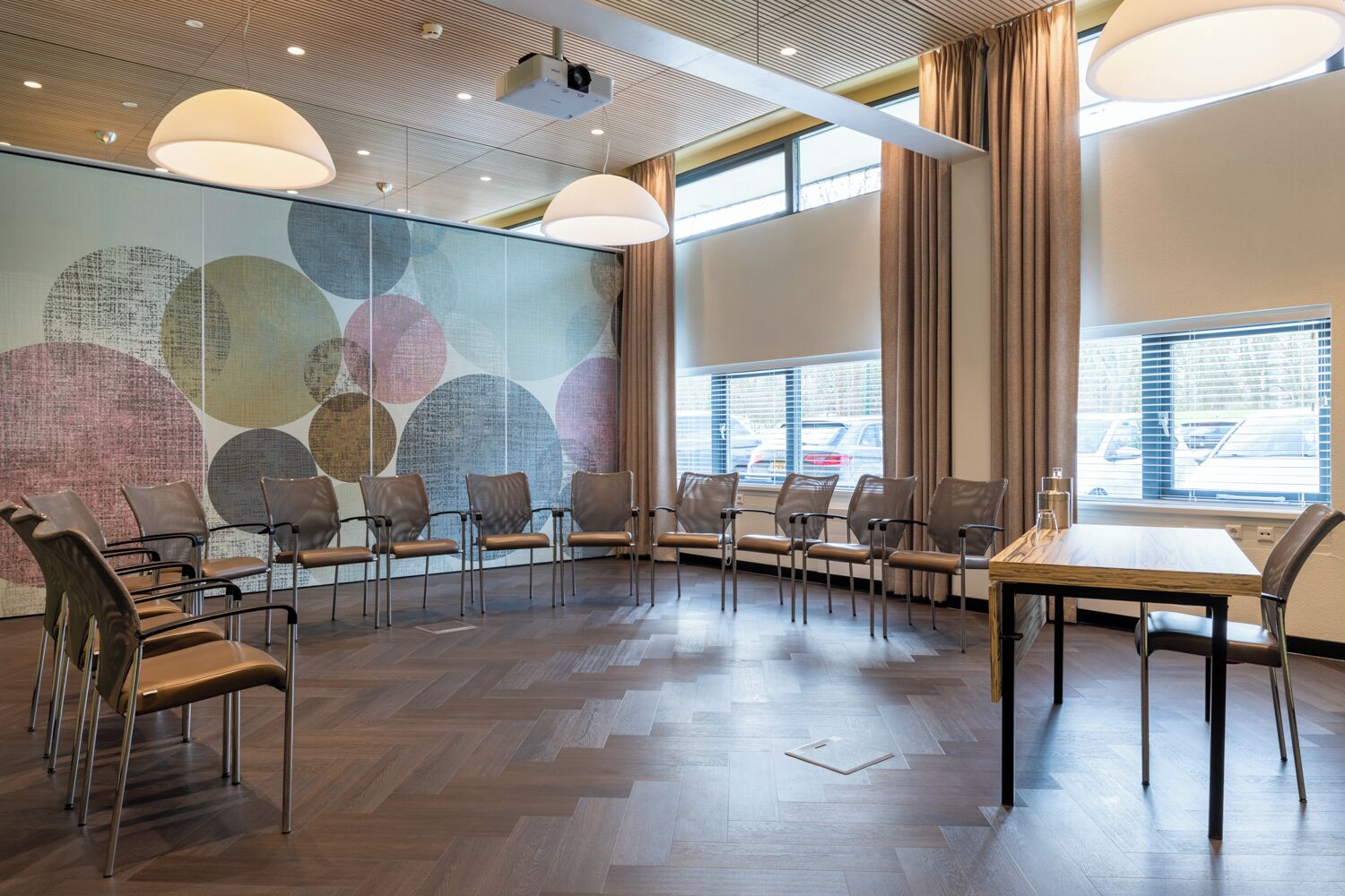 Un salón de conferencias con diseño moderno, que destaca por su suelo de madera en patrón de espiga, sillas de cuero, una mesa de madera y un amplio mural abstracto en la pared.