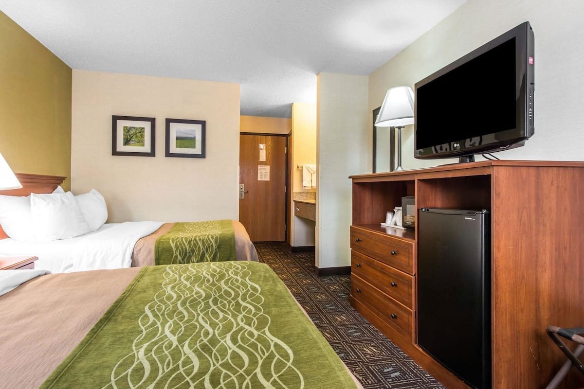 A hotel room with two beds, a TV, a refrigerator, a wooden dresser, and a door leading to a bathroom.