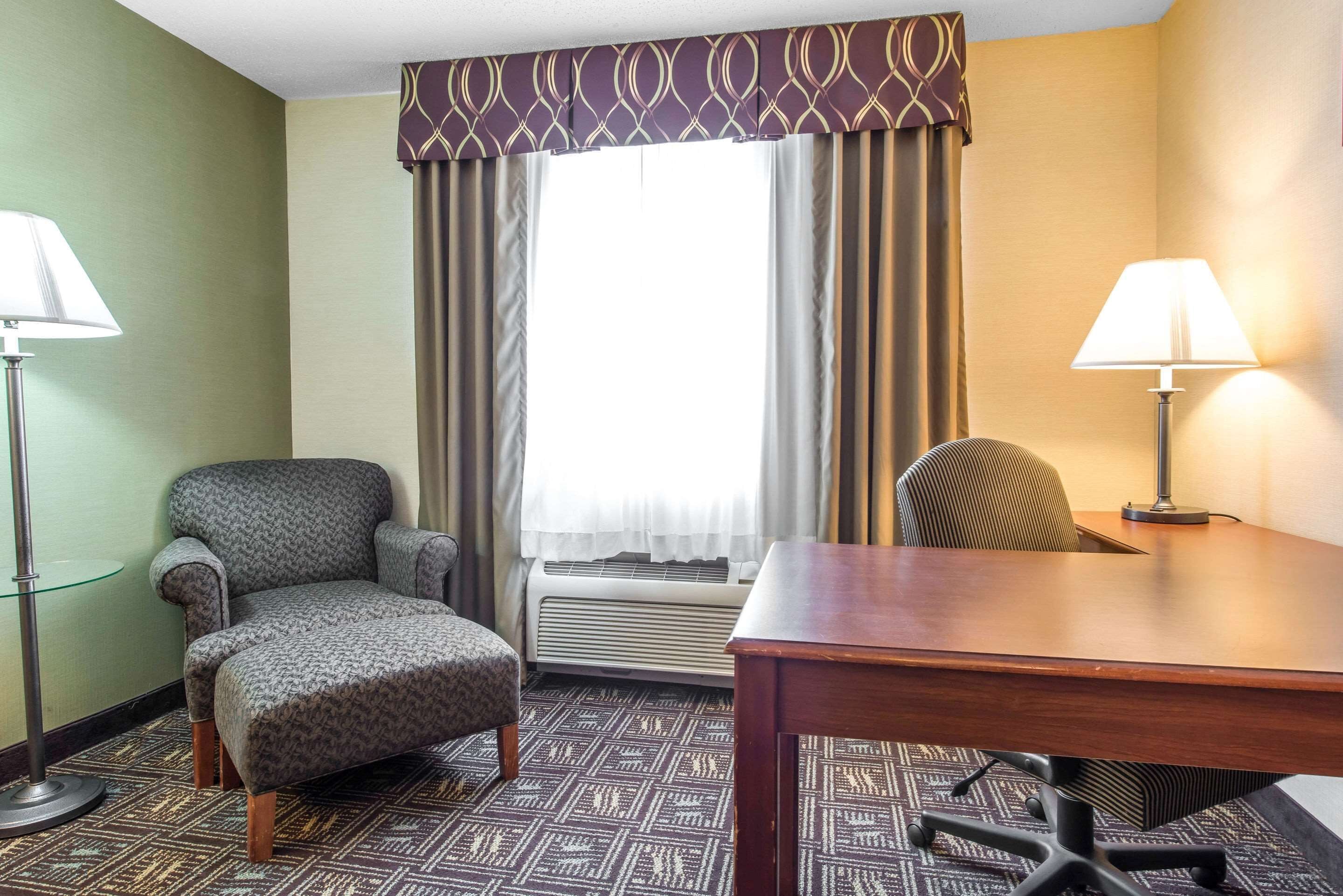 Une chambre d’hôtel avec une moquette à motifs, un bureau en bois, deux chaises, une fenêtre avec des rideaux et deux lampes.