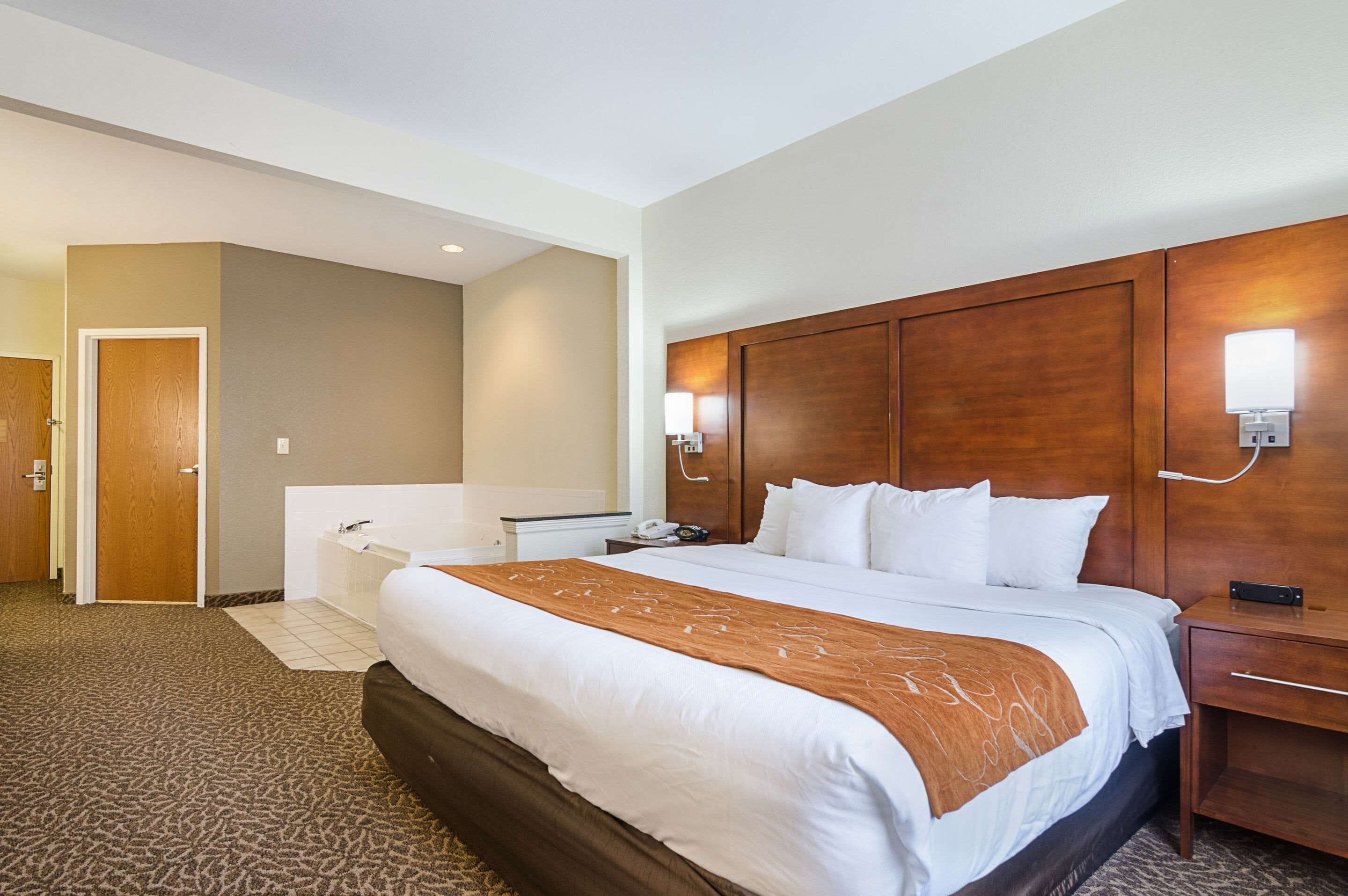 Habitación de hotel con una cama grande, un cabecero de madera, una mesita de noche y un tocador, además de un baño con bañera e inodoro.