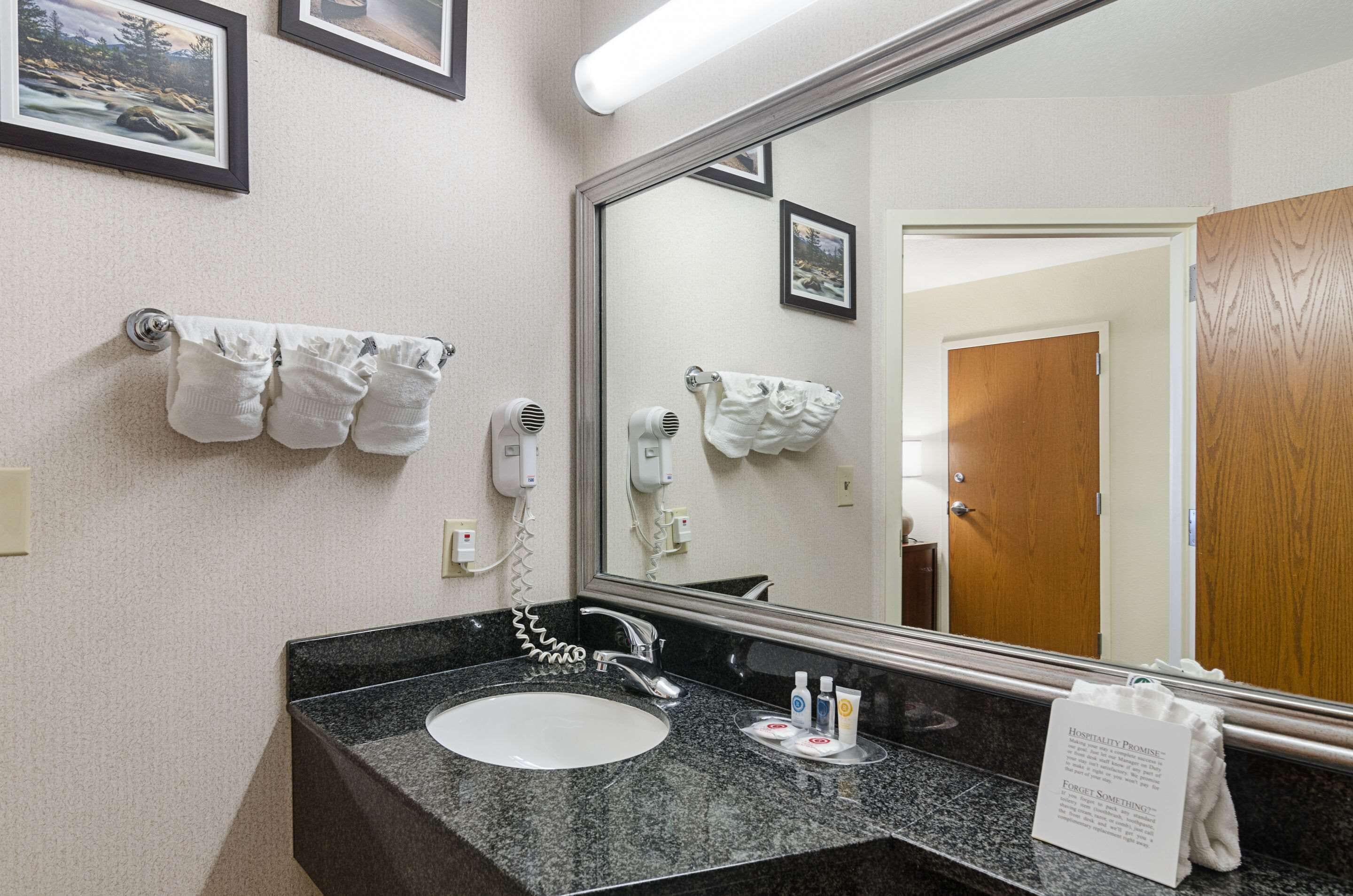A hotel bathroom with a granite countertop sink, a large mirror, and two hair dryers mounted on the wall.
