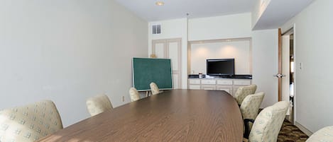 A conference room with a large wooden table, six patterned chairs, a greenboard, a flat-screen TV, and a kitchenette area.