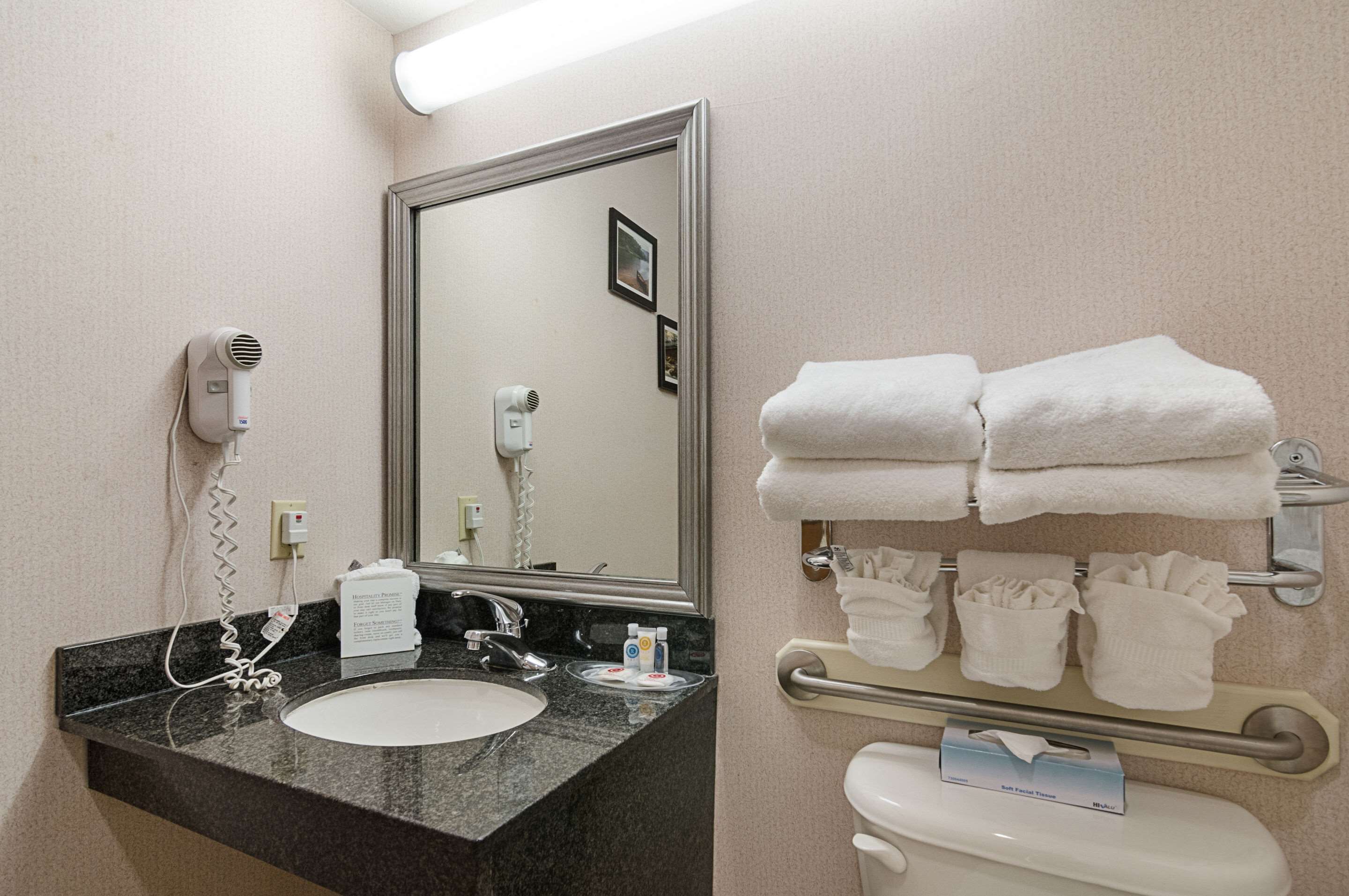 A hotel bathroom with a sink, two hair dryers, a mirror, and a towel rack with folded towels.