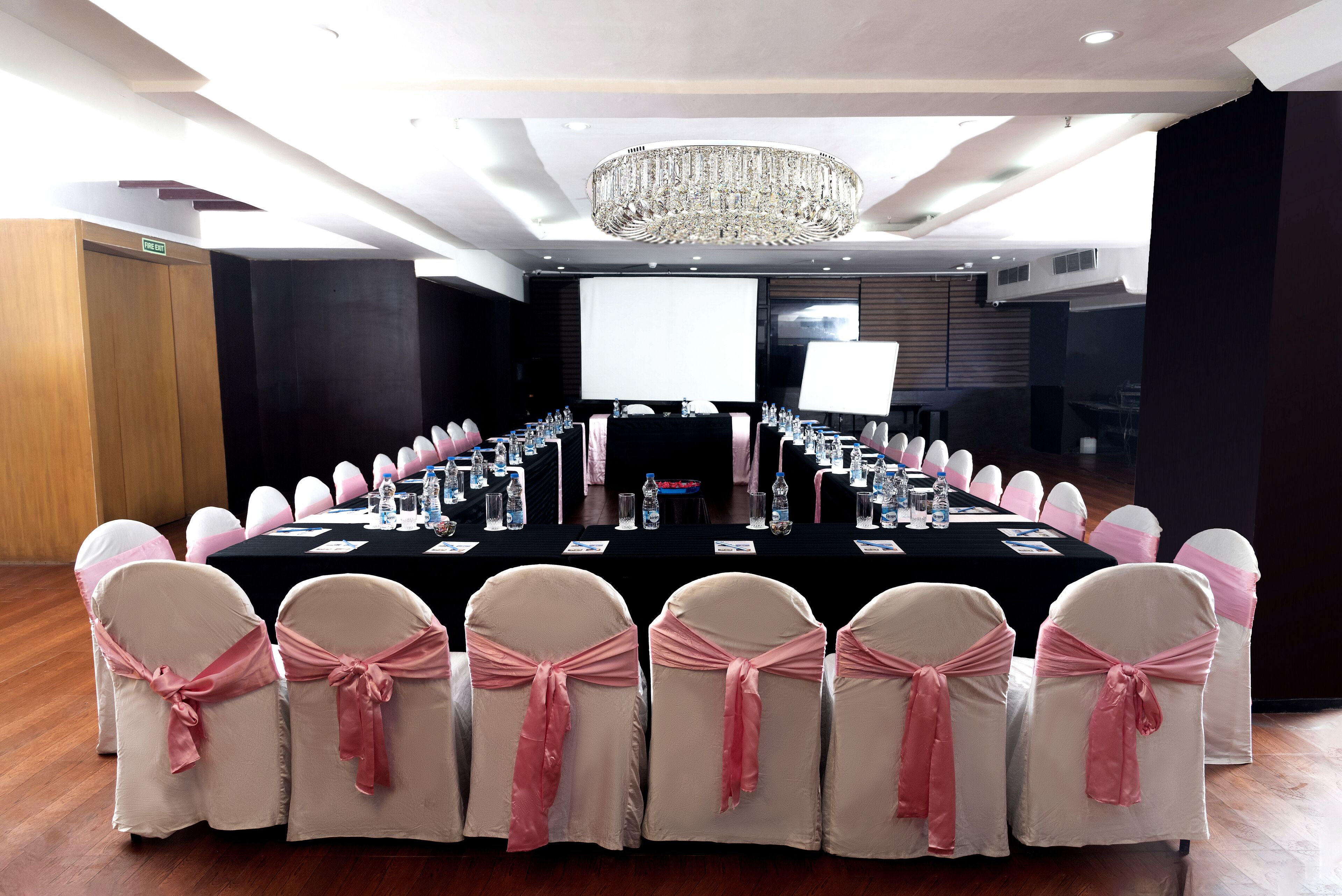 Un salón de conferencias con una larga mesa, sillas adornadas con cintas rosas, una araña y una pantalla grande.