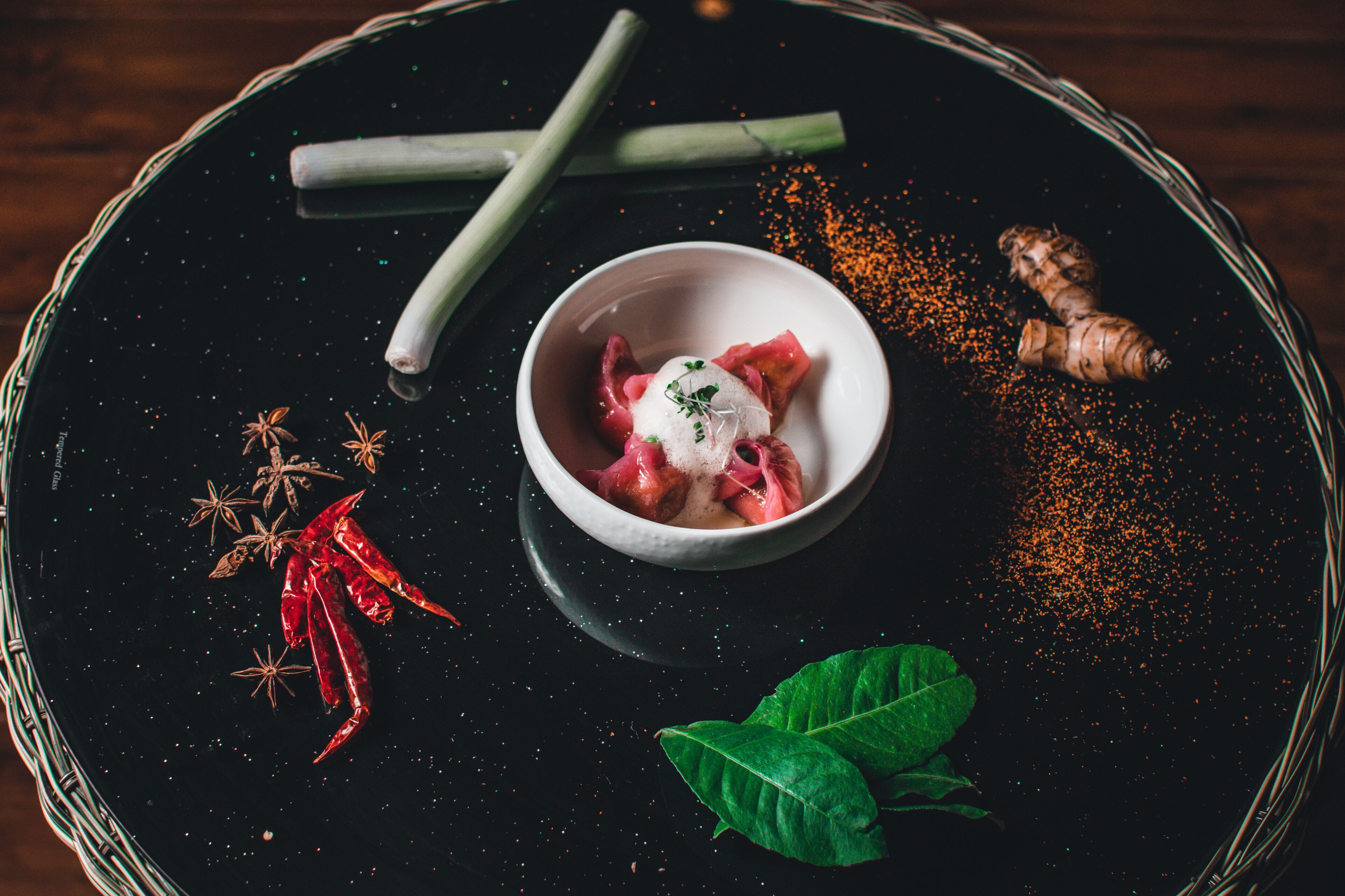 A black tray with a bowl of food, green leaves, red chili peppers, star anise, and a ginger root.