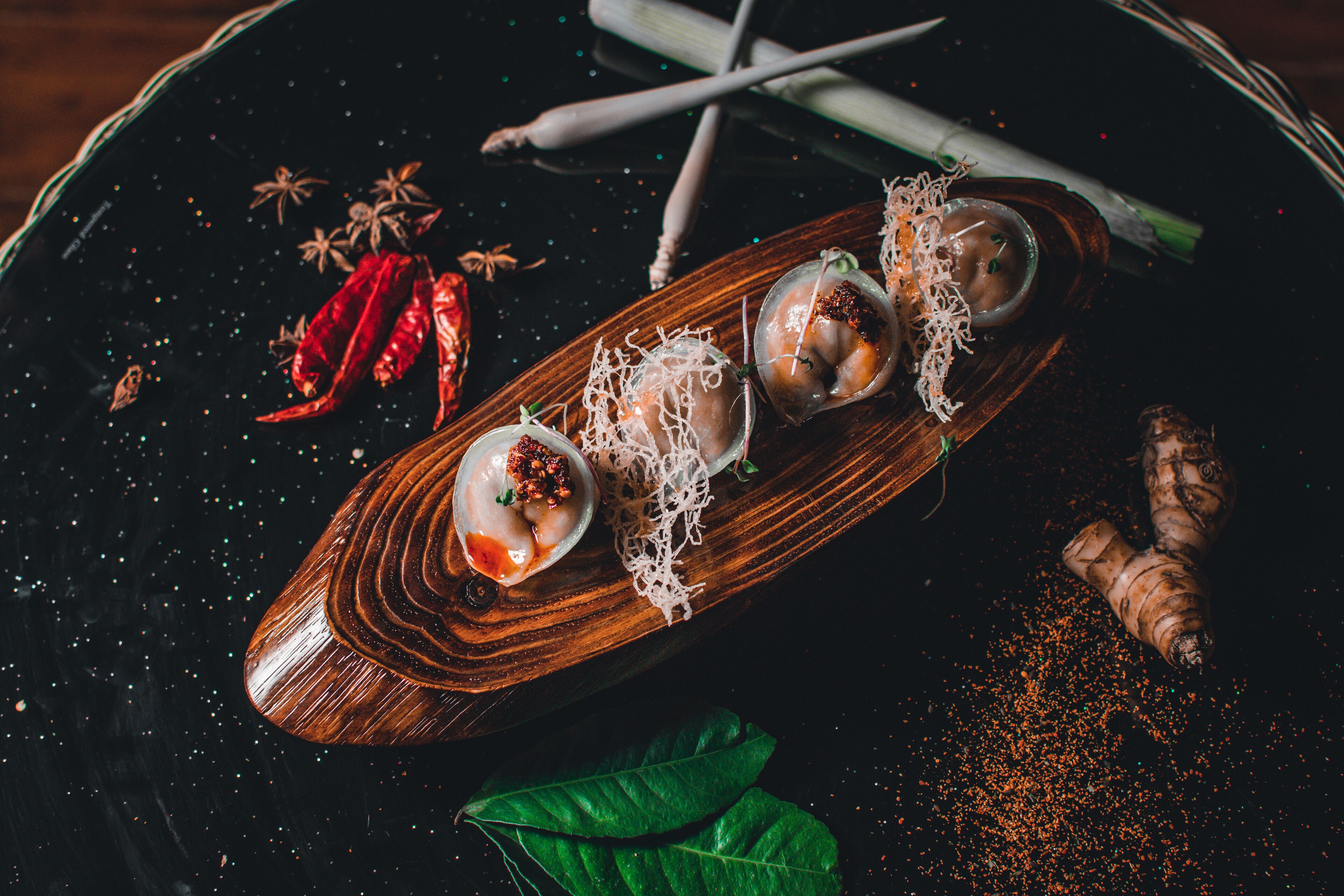 A wooden boat-shaped serving dish with three dumplings, garnished with herbs and spices, on a dark surface with scattered chili peppers and a ginger root.