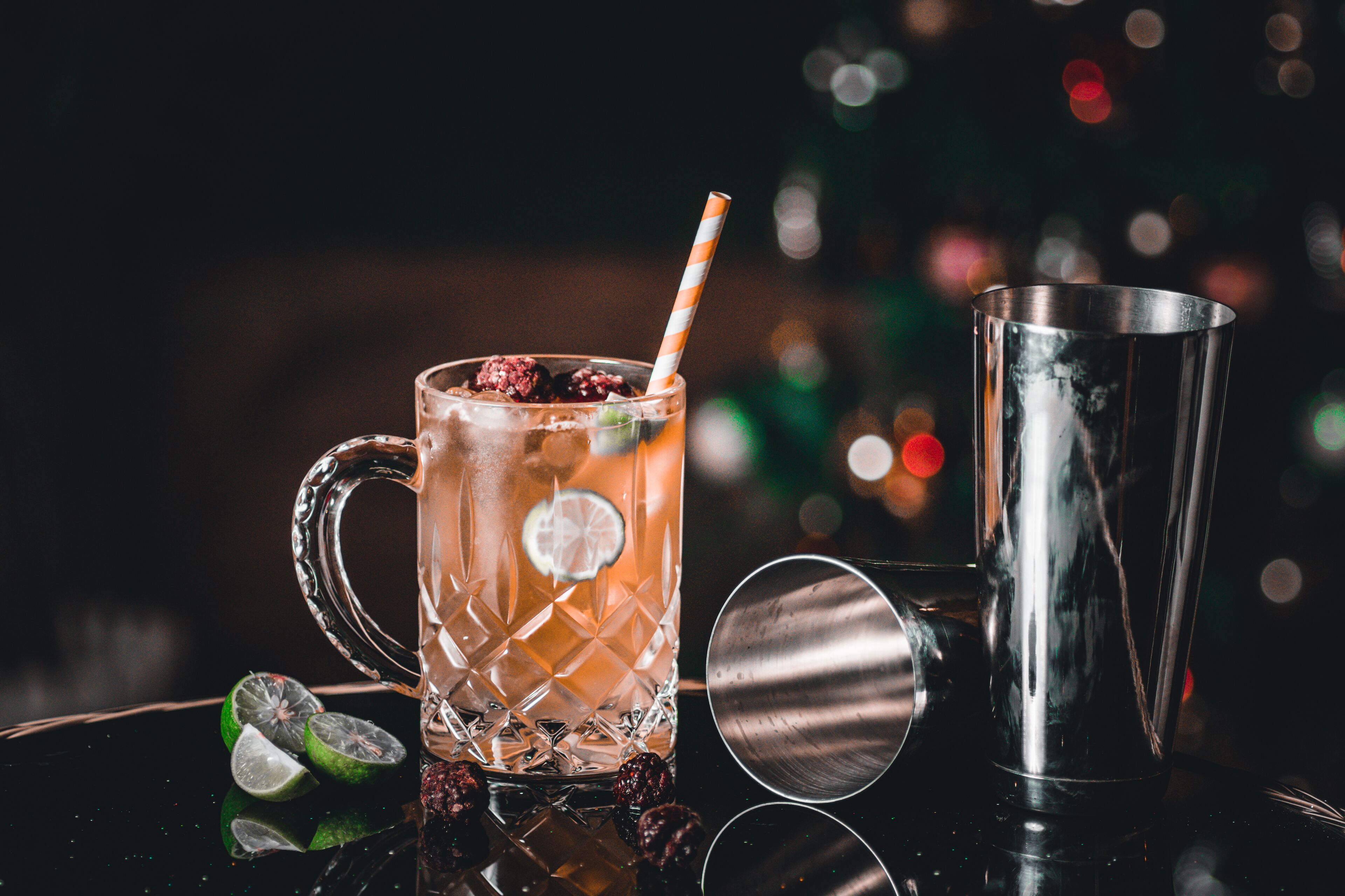 A cocktail glass with a striped straw, ice cubes, and lime slices, accompanied by a metal shaker and lime wedges on a reflective surface.
