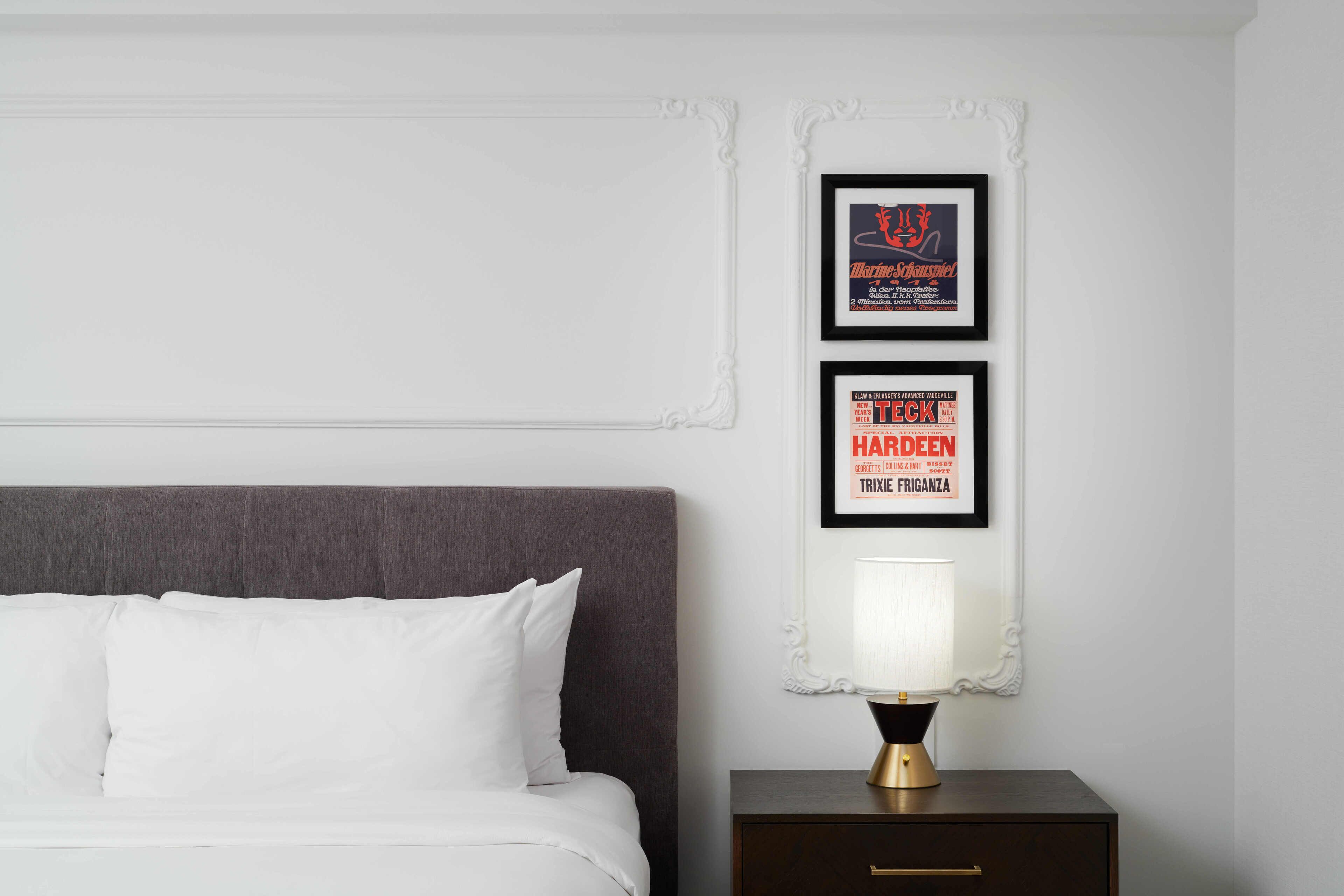 Un lit bien fait, avec du linge de lit blanc, une tête de lit sombre et une table de chevet sur laquelle se trouve une lampe. Deux affiches encadrées sont accrochées au mur au-dessus du lit.