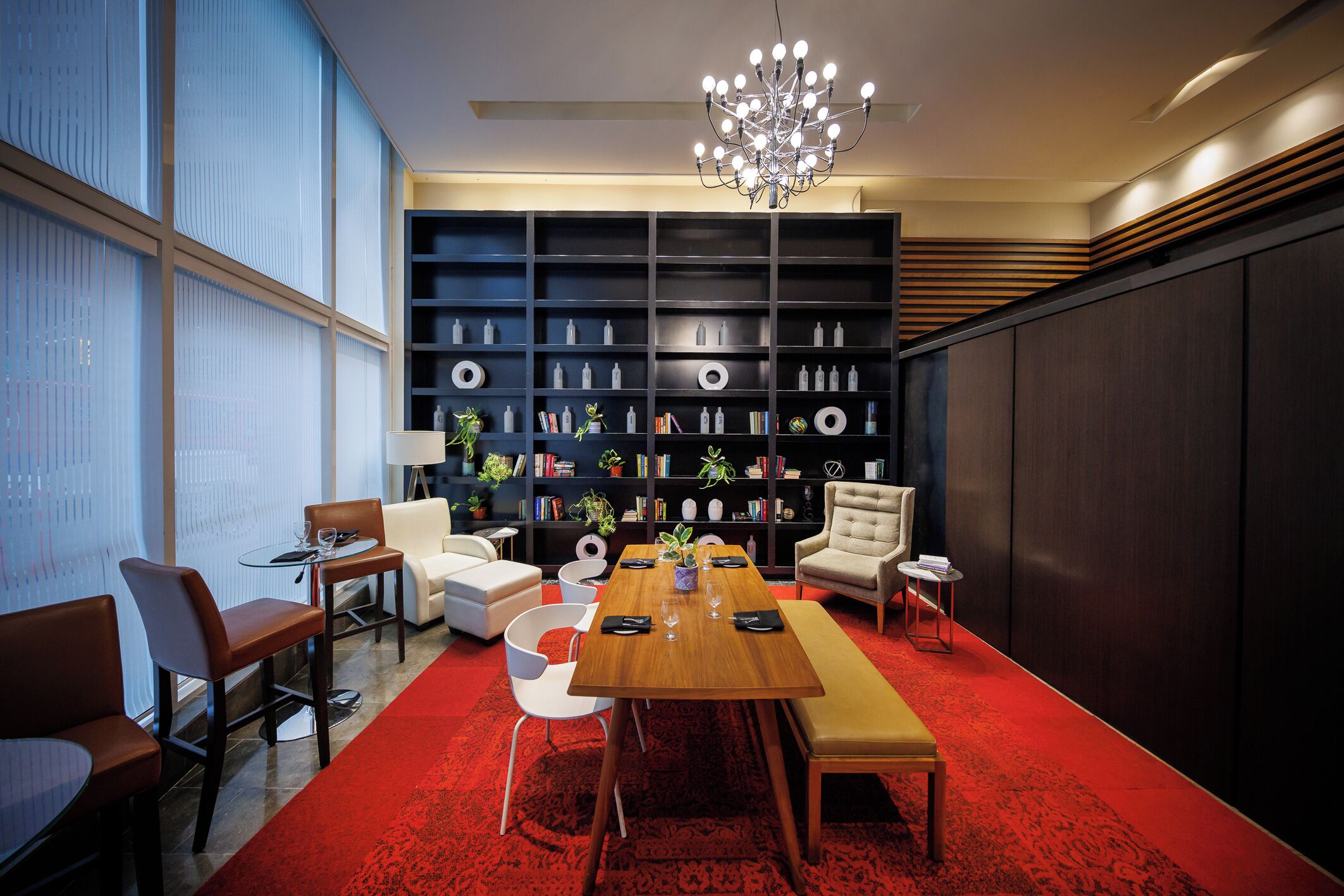 A modern dining area with a wooden table, chairs, and a red carpet. There is a bookshelf with books and decorative items, and a chandelier above.
