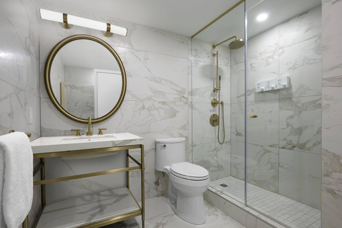 A modern bathroom with a marble floor, a gold-framed mirror, a white sink, a toilet, and a glass-enclosed shower area.