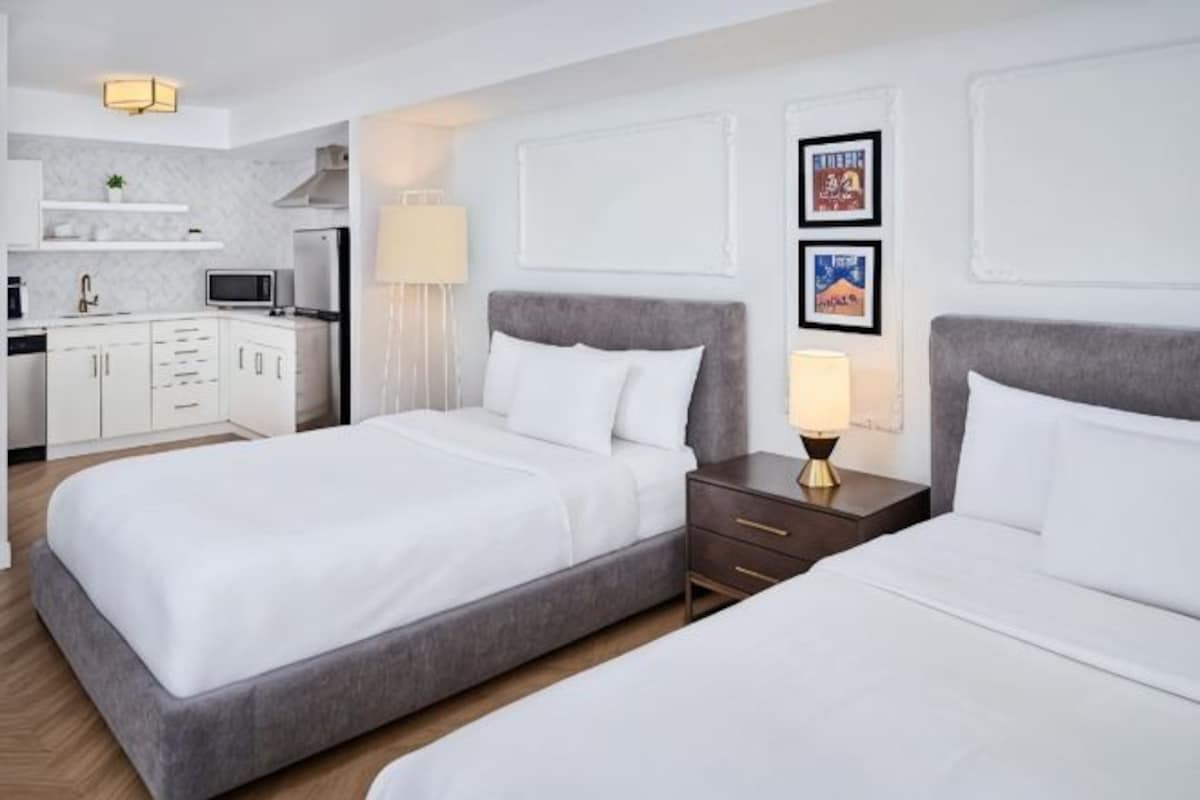 Habitación de hotel con dos camas, una kitchenette pequeña y un televisor montado en la pared.