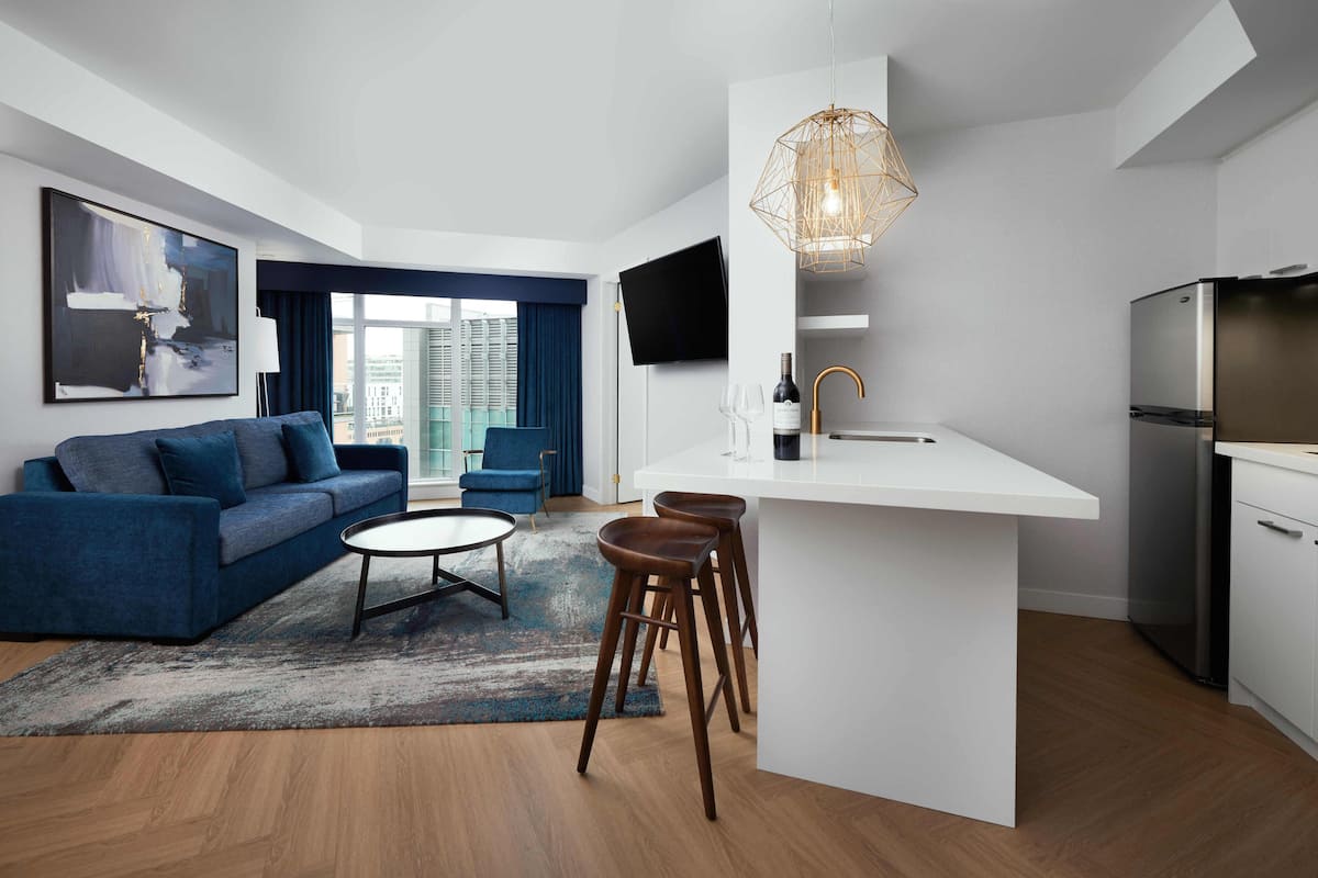 A modern living room with a blue sofa, a coffee table, and a kitchen area with a white countertop and bar stools.
