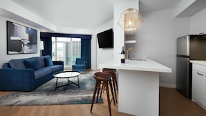 A modern living room with a blue sofa, a coffee table, and a kitchen area with a white countertop and bar stools.