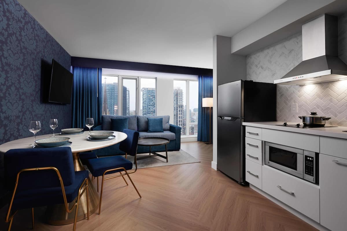A modern kitchen with a built-in microwave, a refrigerator, and a dining area with a round table and blue chairs.