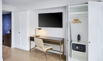 Een moderne hotelkamer met een houten bureau, een comfortabele stoel, een flatscreen televisie aan de muur, een kledingkast met ophanging en een klein safe.