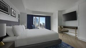A modern hotel room with a large bed, a desk, a chair, a flat-screen TV, and a view of the city skyline.