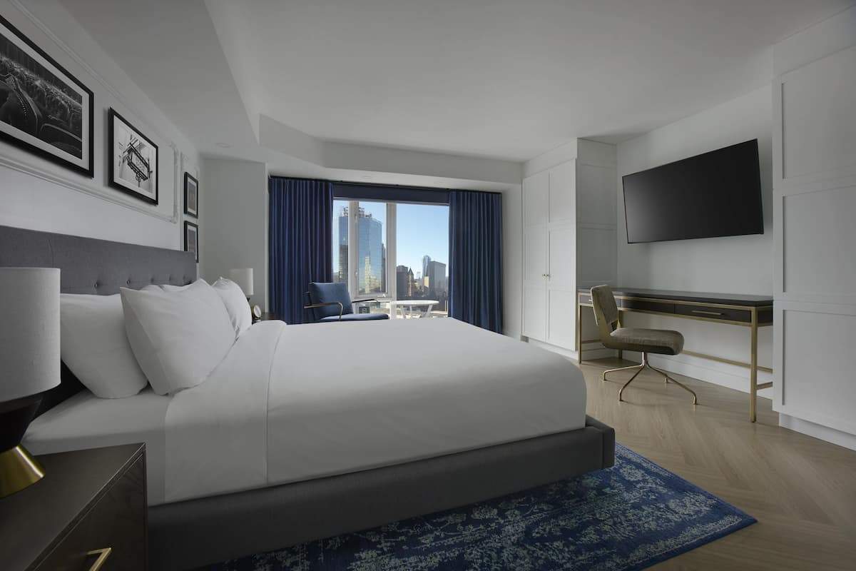 A modern hotel room with a large bed, a desk, a chair, a flat-screen TV, and a view of the city skyline.