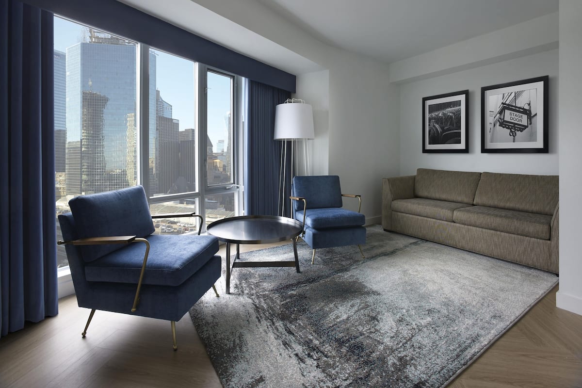 A modern hotel room with a large window offering a city view, a round coffee table, two armchairs, a sofa, and two framed pictures on the wall.