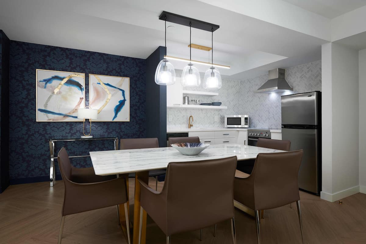 A modern kitchen with a marble countertop, stainless steel appliances, and a dining area with a white table and brown chairs.