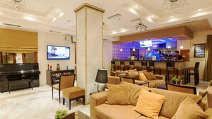 A hotel lobby with a piano, comfortable seating, a bar area, and a television displaying a news program. - Venus Hotel & Suites (Zakynthos)