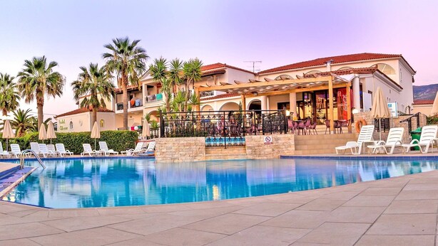 A hotel with a pool, lounge chairs, palm trees, and a restaurant. - Venus Hotel & Suites (Zakynthos)