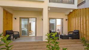 A covered outdoor area with wooden flooring, two seating areas with wicker furniture, and a glass door leading to an interior space. - Venus Hotel & Suites (Zakynthos)