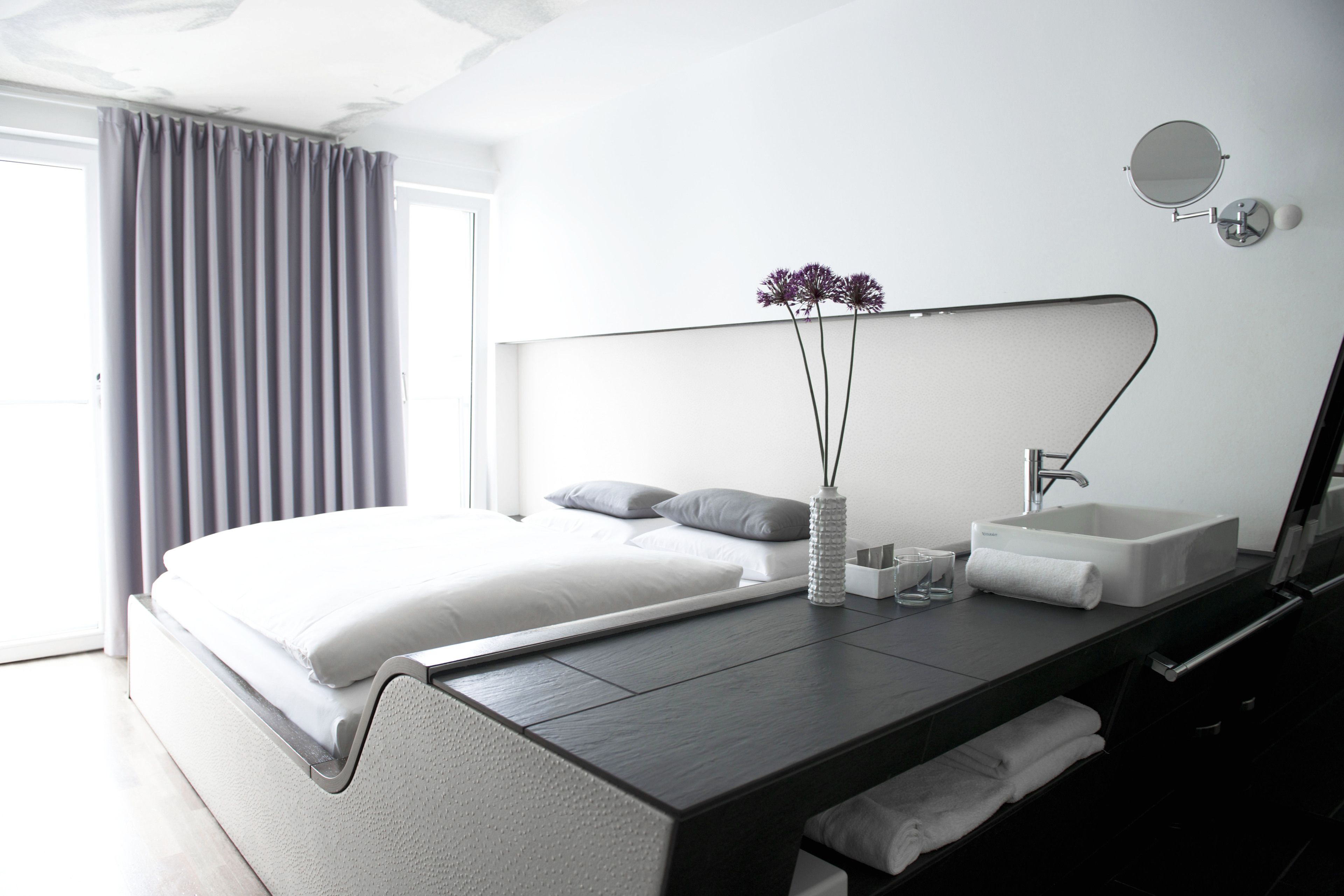 Una habitación de hotel moderna con una cama grande, una mesita de noche con un florero, un espejo y un lavabo.